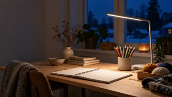 Ett städat hobby- och läsbord på kvällen där en omärkt smal skrivbordslampa på arm ger jämnt vitt ljus över färgpennor, garn och ett tomt skissblock