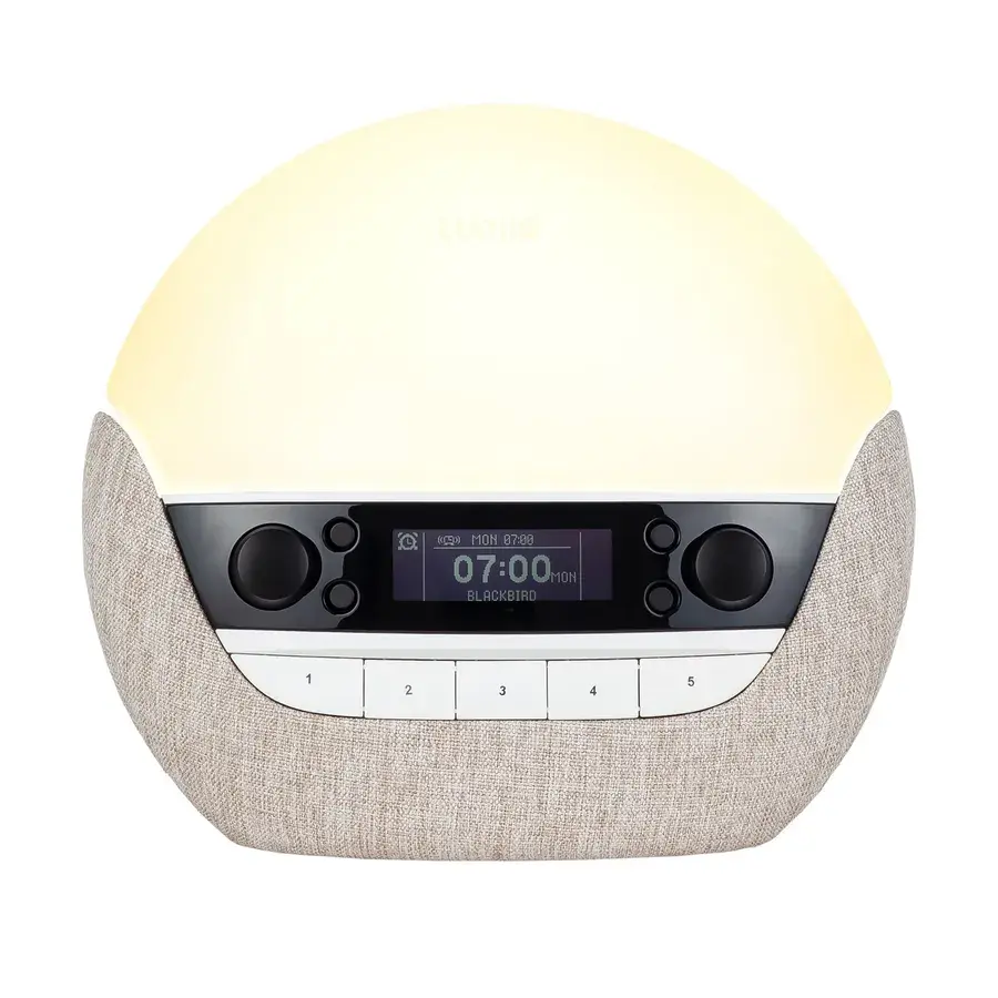 Lumie Bodyclock Luxe FM, en stor kupolformad väckningslampa med beige textilsockel, knappar och display, fotad mot vit bakgrund